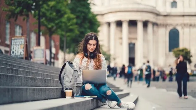 Una chica contenta, sentada con una laptop sobre unas escaleras en la calle.